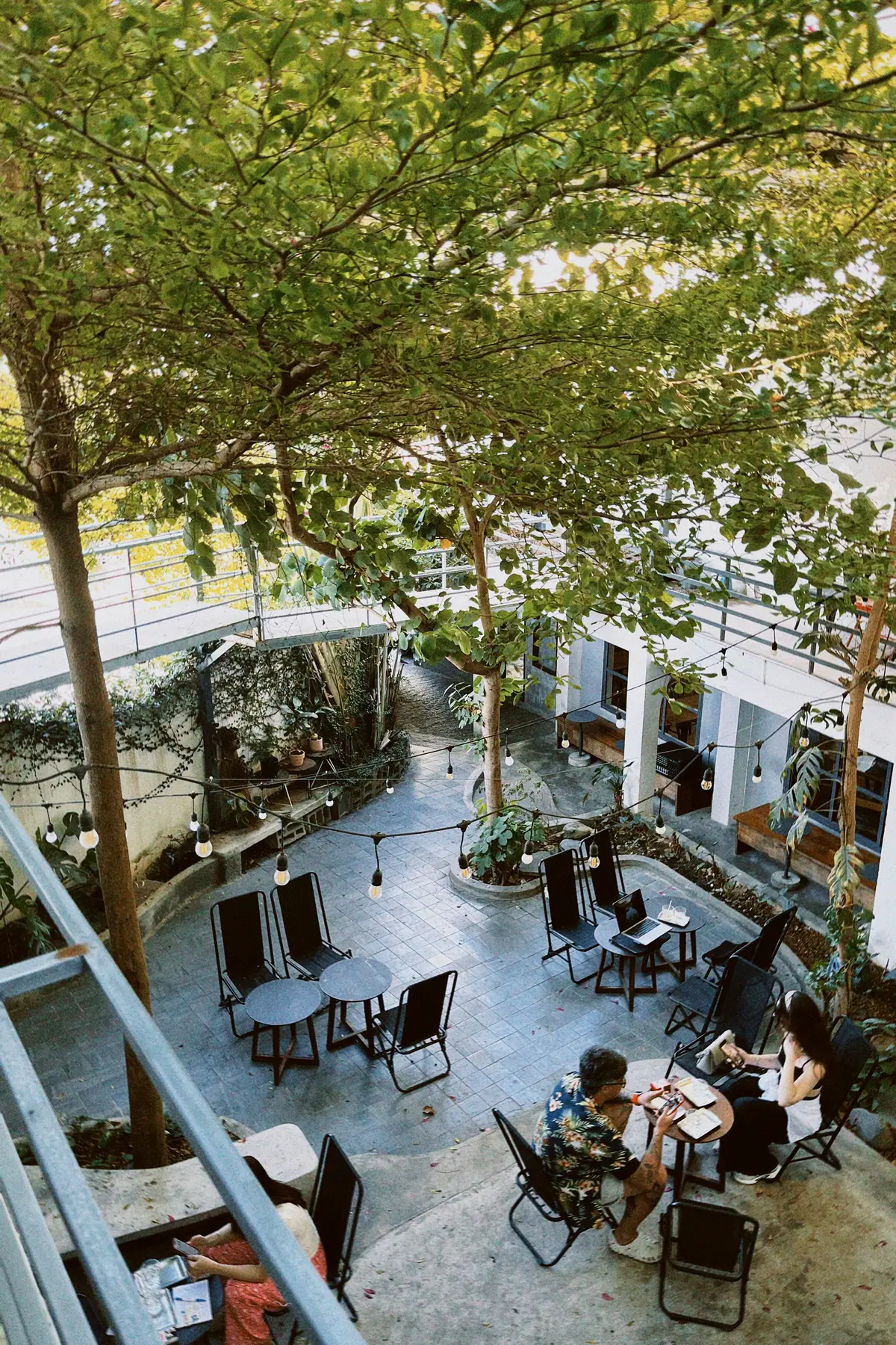 Courtyard cafe with trees, string lights and metal chairs seen from a balcony