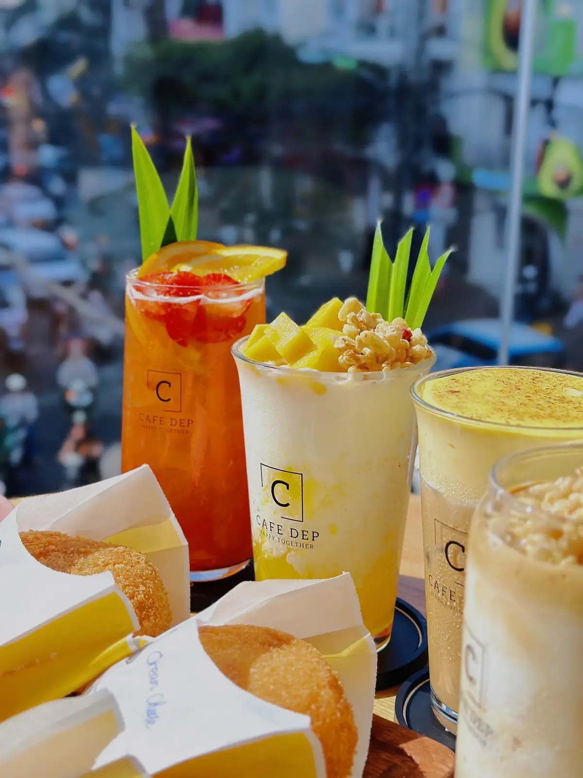 Three tall glasses of orange tea, mango drink and a creamy yellow drink with a snack beside them