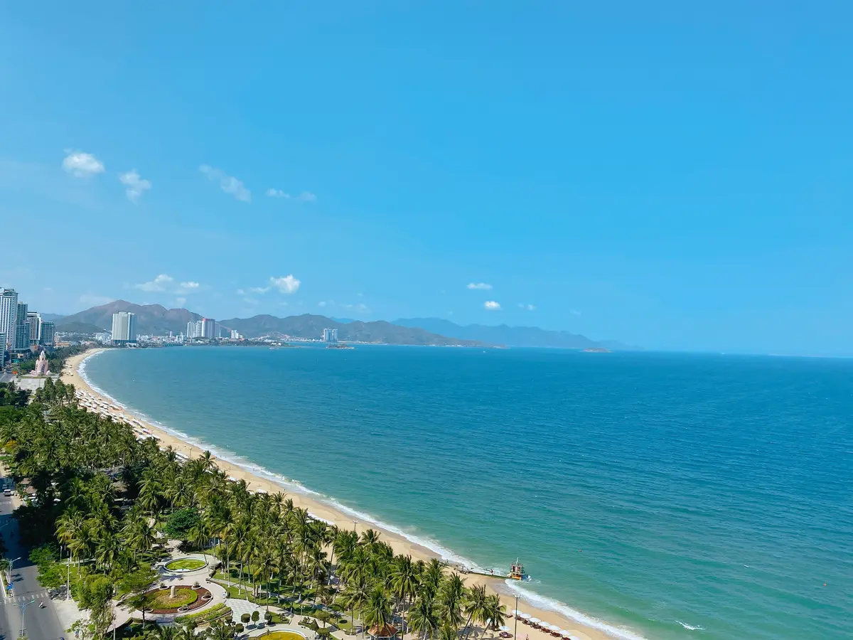 Long curve of Nha Trang beach and palm-lined park seen from above, with calm blue sea, clear sky and mountains across the bay