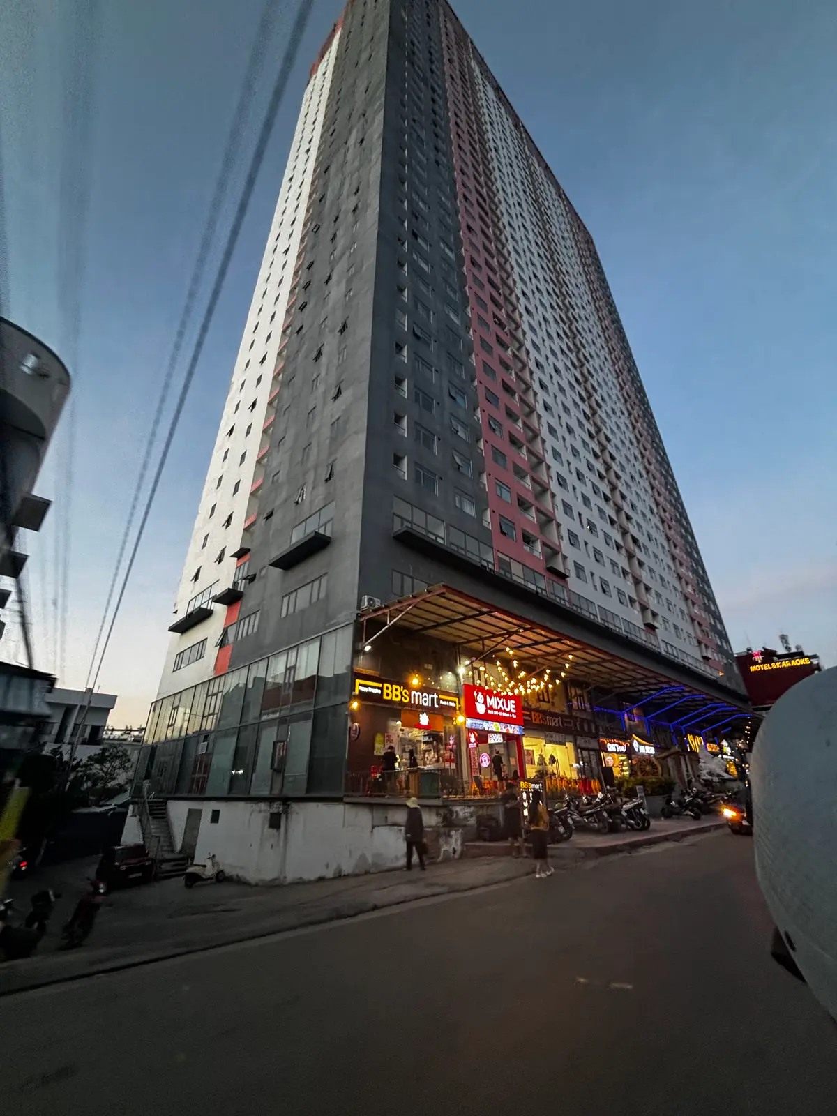 Apartment tower seen from the street at dusk with shops and signs lit along its ground floor