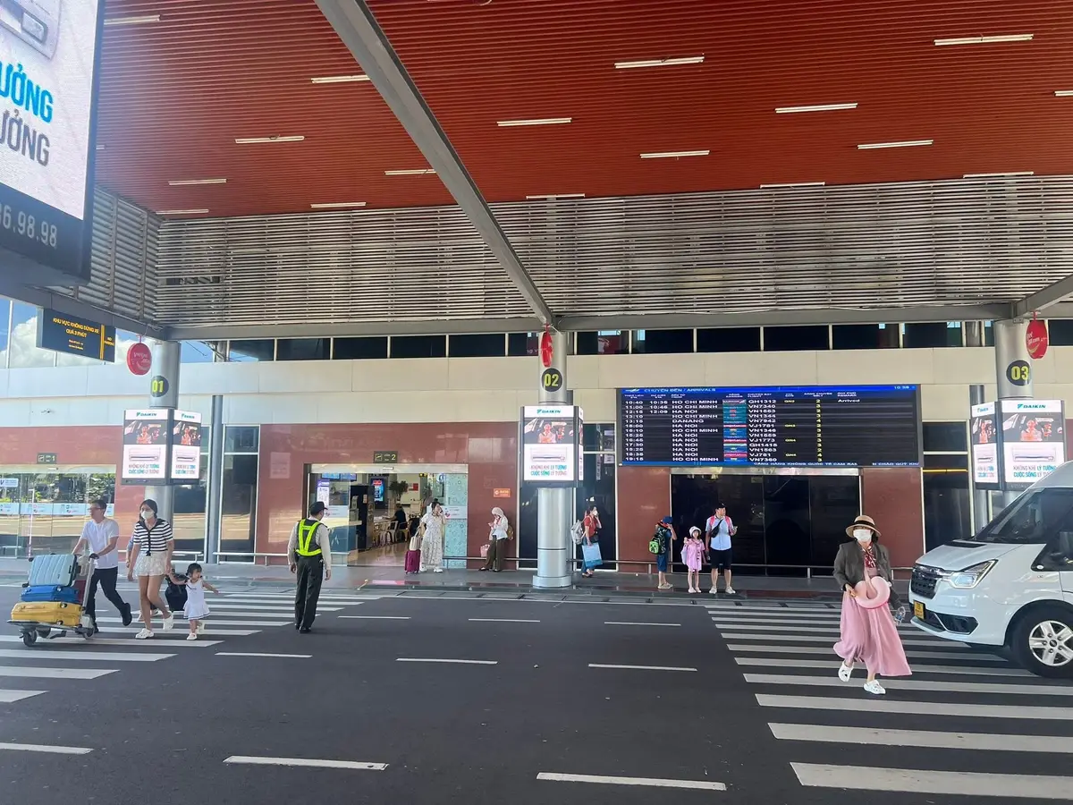 Pedestrian crossing in front of the arrivals entrance at Cam Ranh International Airport with a white car, passengers with luggage and a covered forecourt