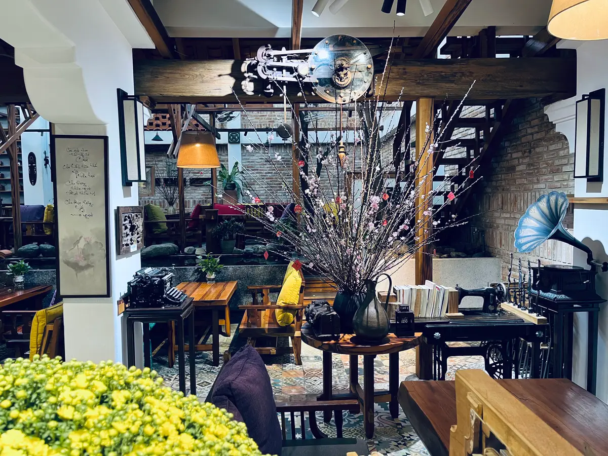 Dining room with wooden tables, a gramophone, framed pictures and an open staircase, with yellow flowers in front
