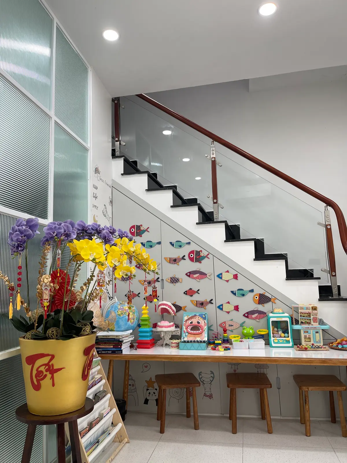 Waiting-area corner under a staircase with a low table, stools, books and orange flowers