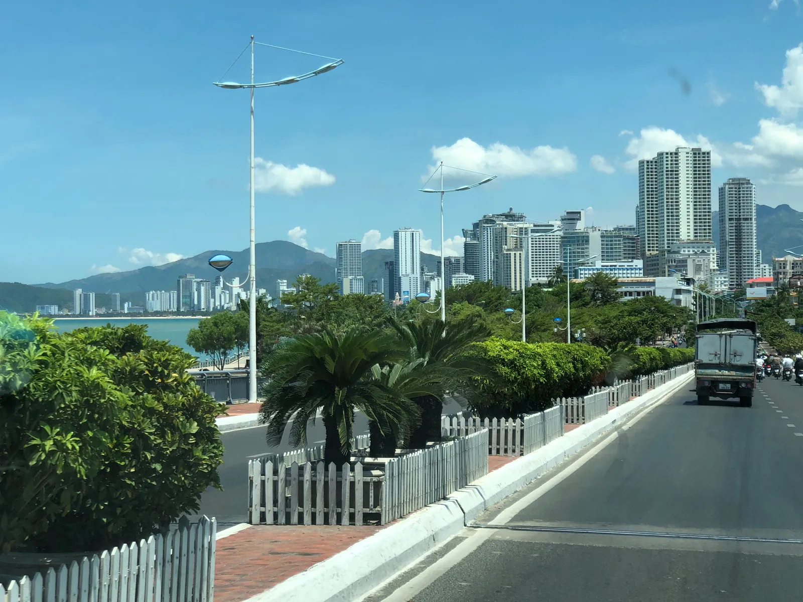 Seaside road in Nha Trang with palm trees, a white railing, high-rise buildings and mountains in the distance