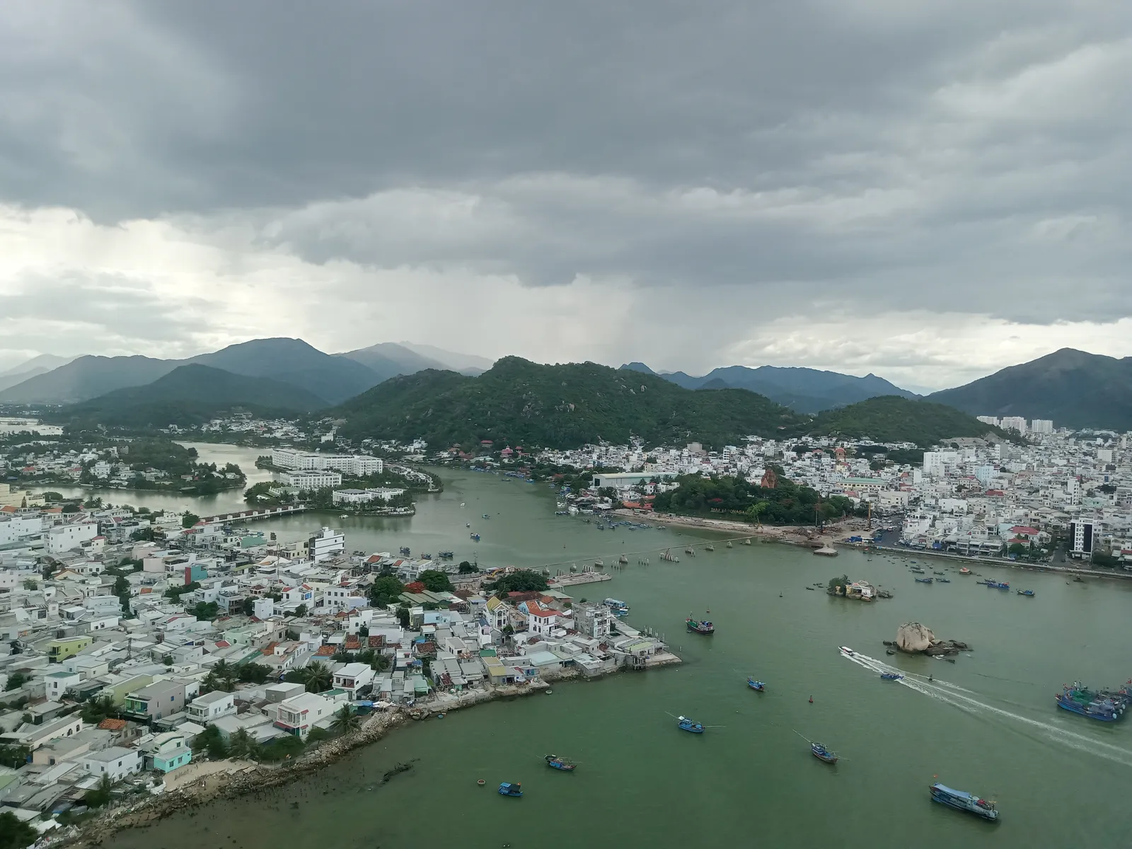 Aerial view of Nha Trang city under a cloudy sky, with the river, boats, dense housing and green hills in the distance