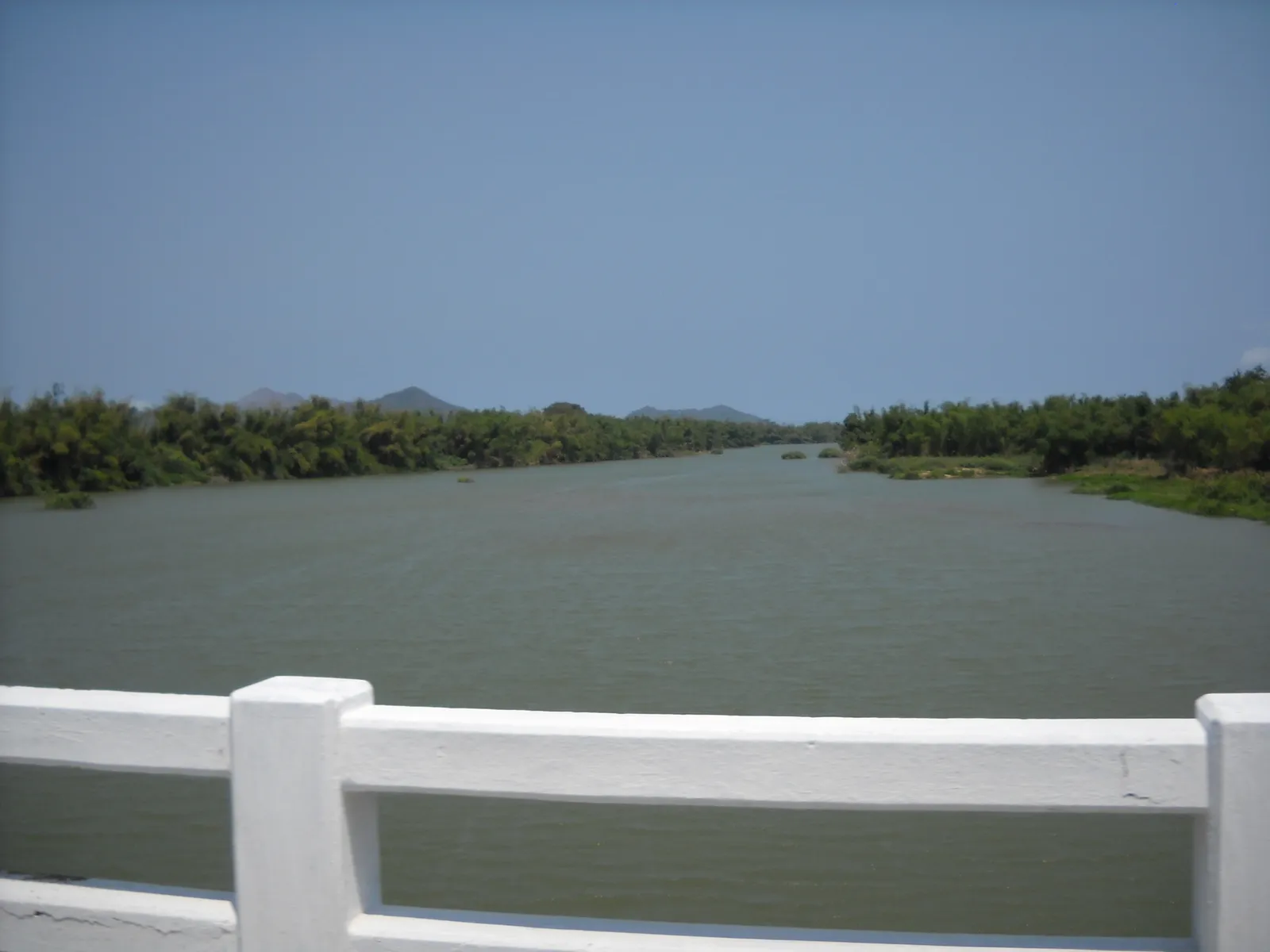 Wide brown river with a white bridge railing in the foreground and low green hills behind