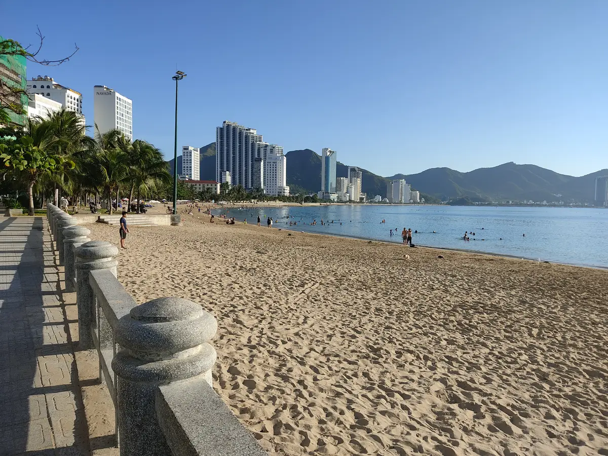 Sandy beach beside a white stone balustrade and a paved promenade, with high-rise buildings and mountains in the distance