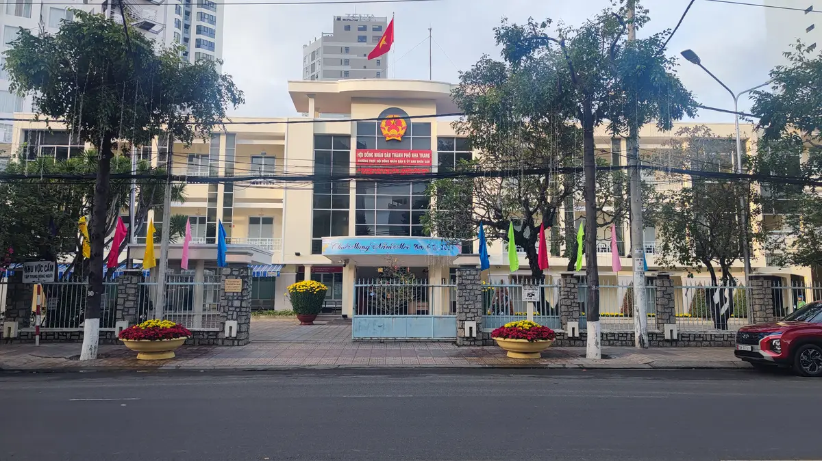 Cream government building with a coat of arms above the entrance, flags in front and trees along the street.