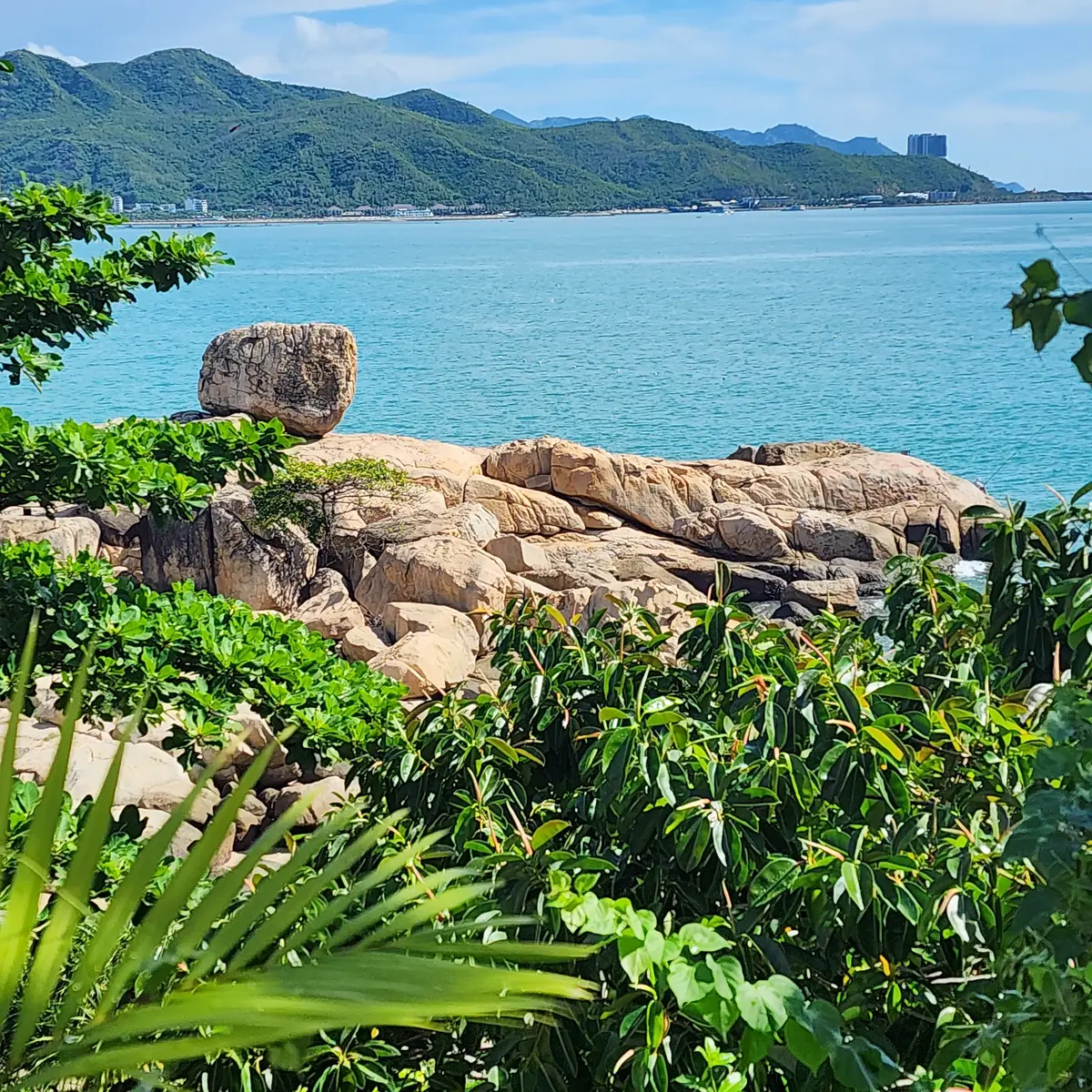 Large balanced boulder on a rock ledge with green plants and turquoise sea