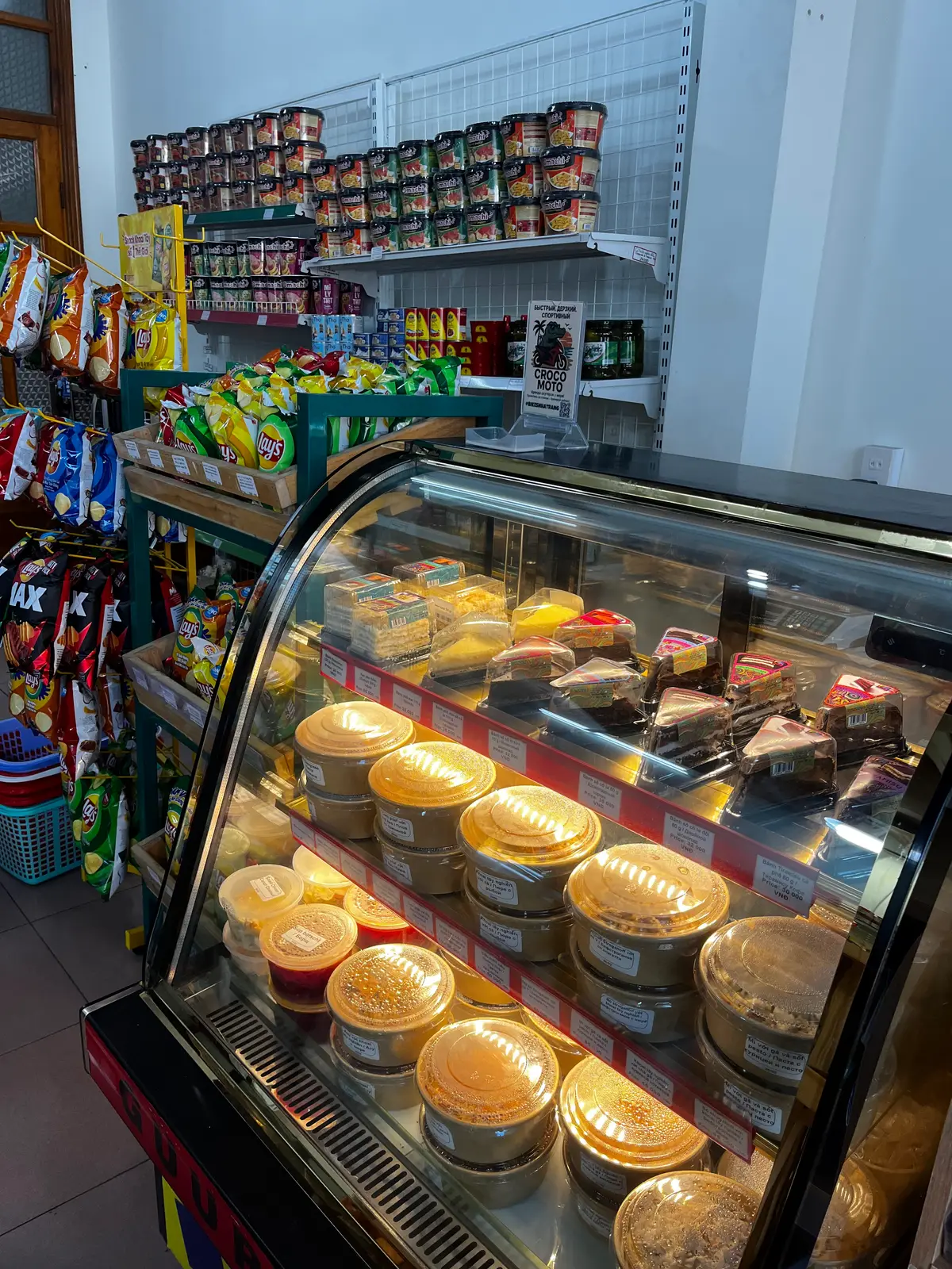 Glass deli case of ready-made dishes in round trays beside shelves of crisps