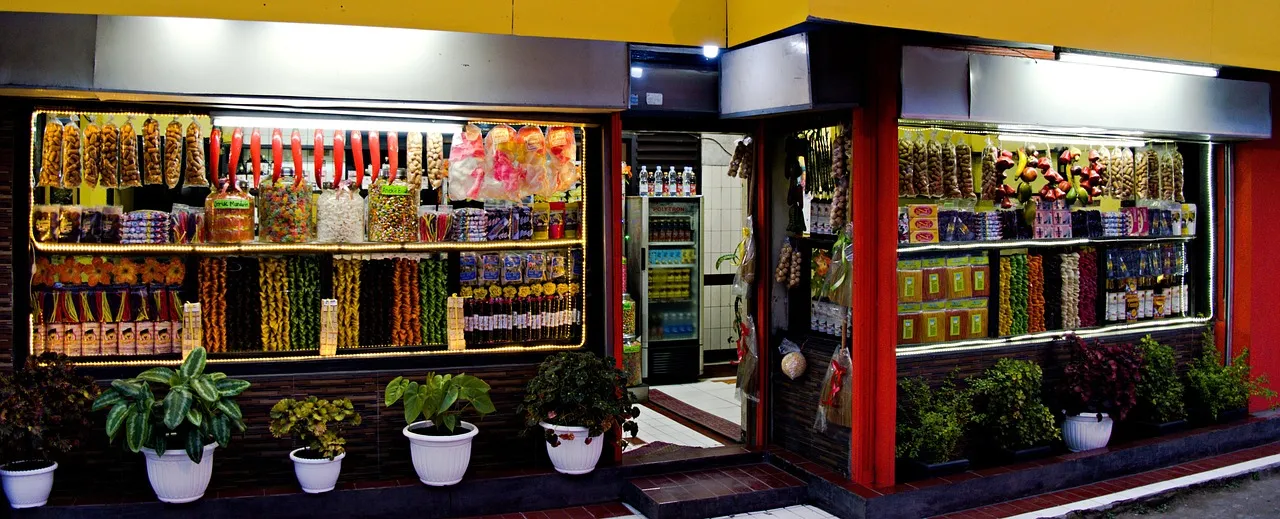Lit corner shop front with jars of snacks and sweets in the windows and potted plants outside