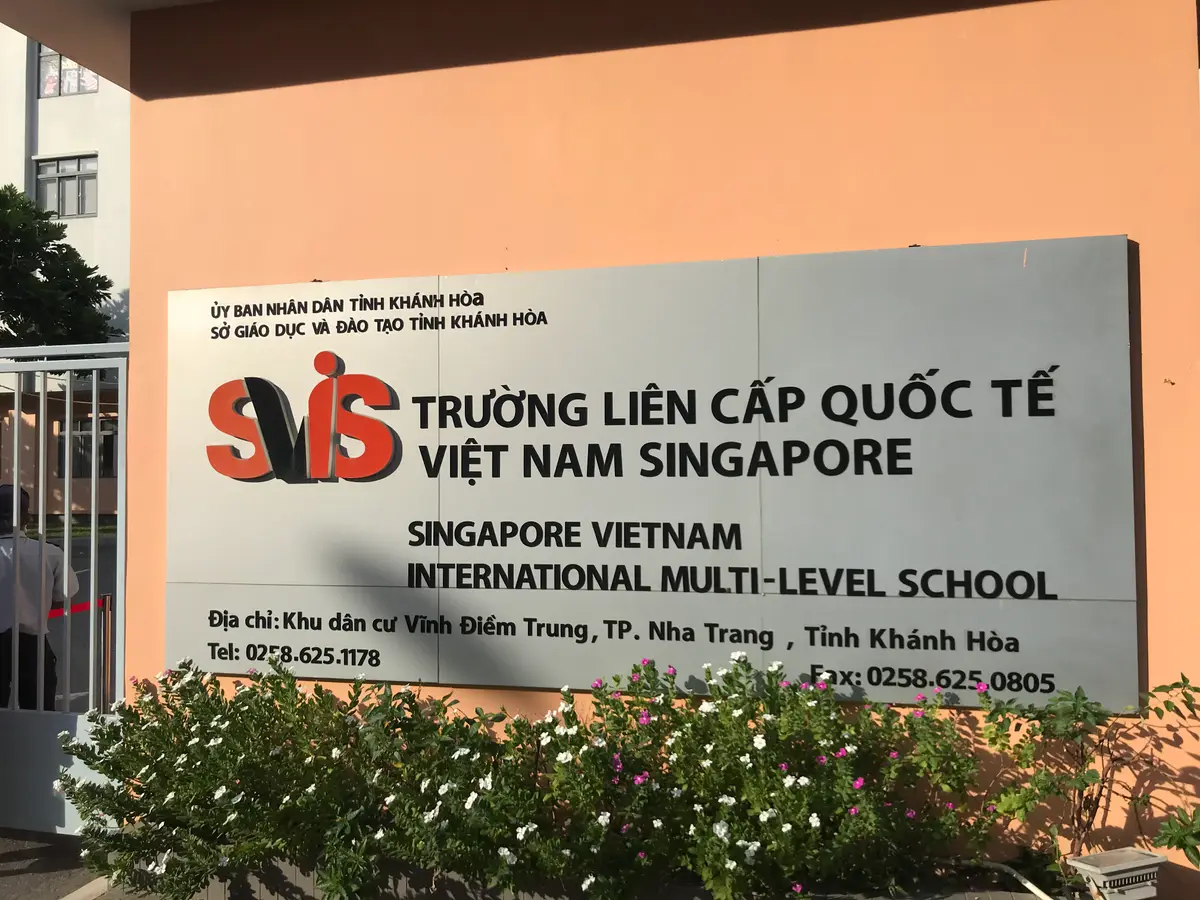 School name plate in English and Vietnamese on an orange wall behind shrubs