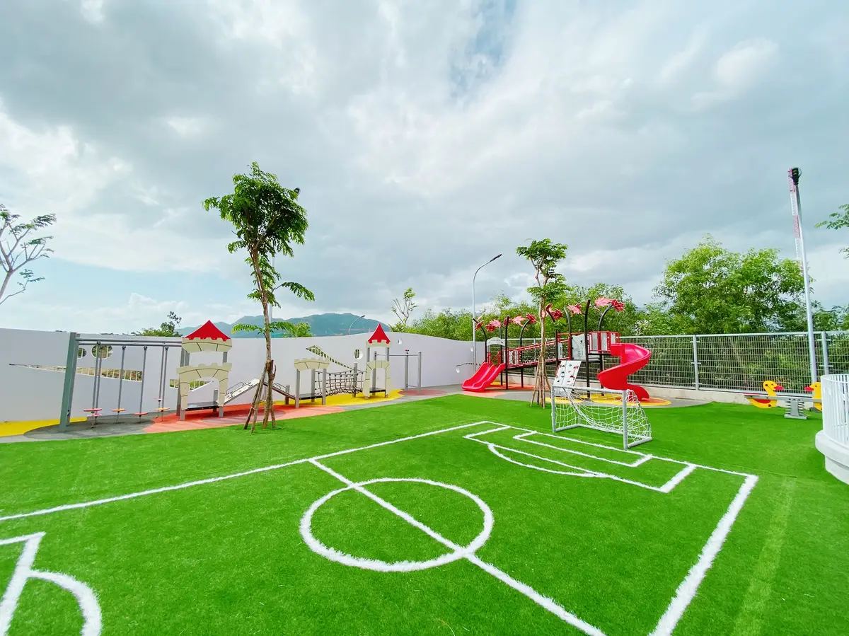 A playground with a green artificial-turf football pitch, climbing frames and slides behind a white wall.