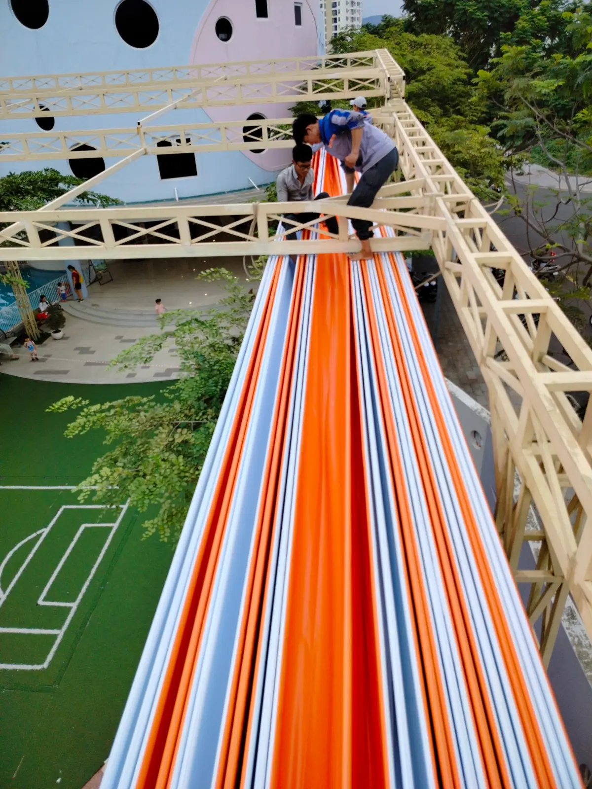 A top-down view of a long orange and white slide beside a wooden frame above a green pitch.
