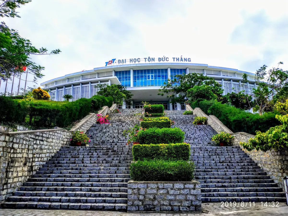 A wide white campus building with a blue glass front above a long stone staircase and trimmed hedges.