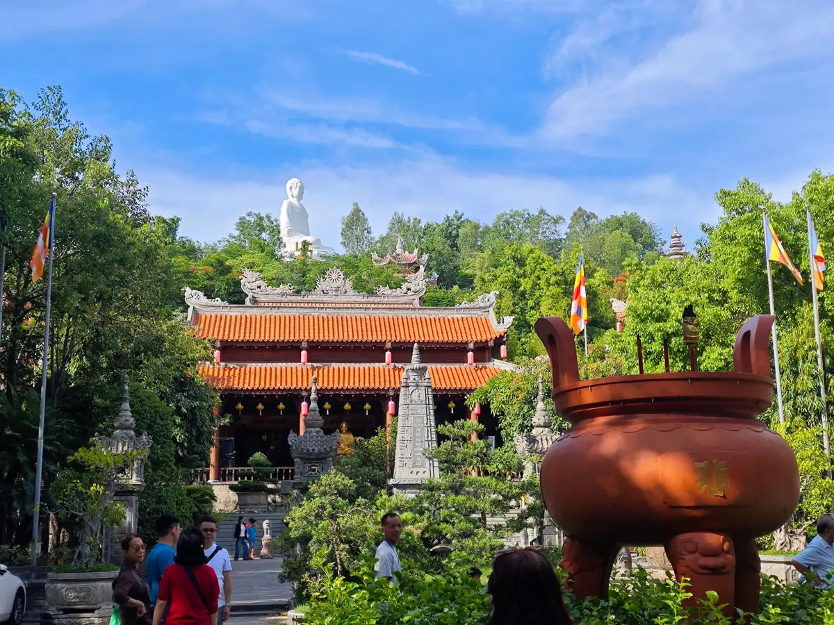 Large bronze incense burner in front of the red tiled hall with the white Buddha statue on the hill