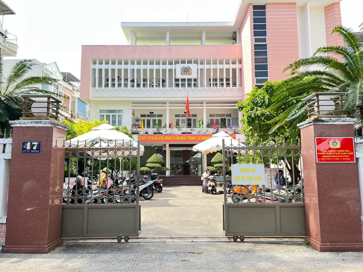 Pink government building behind a metal gate, with a street number plate on the gate pillar, a red police sign and a parasol in the yard.