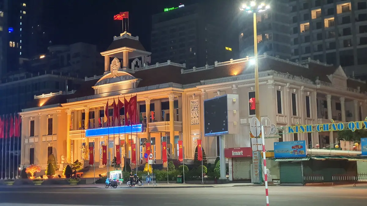 Cream colonial-style government building with a central tower, lit up at night with flags along the front, seen from across the street.
