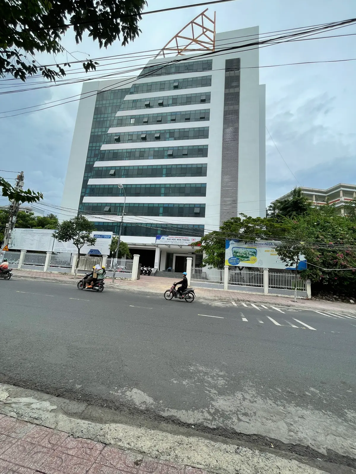 Tall blue-glass university building behind a low wall and a signboard, with motorbikes riding past on the street