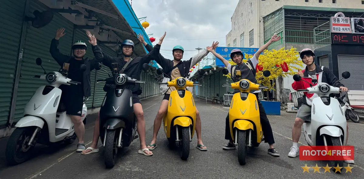 Three riders in helmets on two yellow and one black electric scooters in a street, arms raised