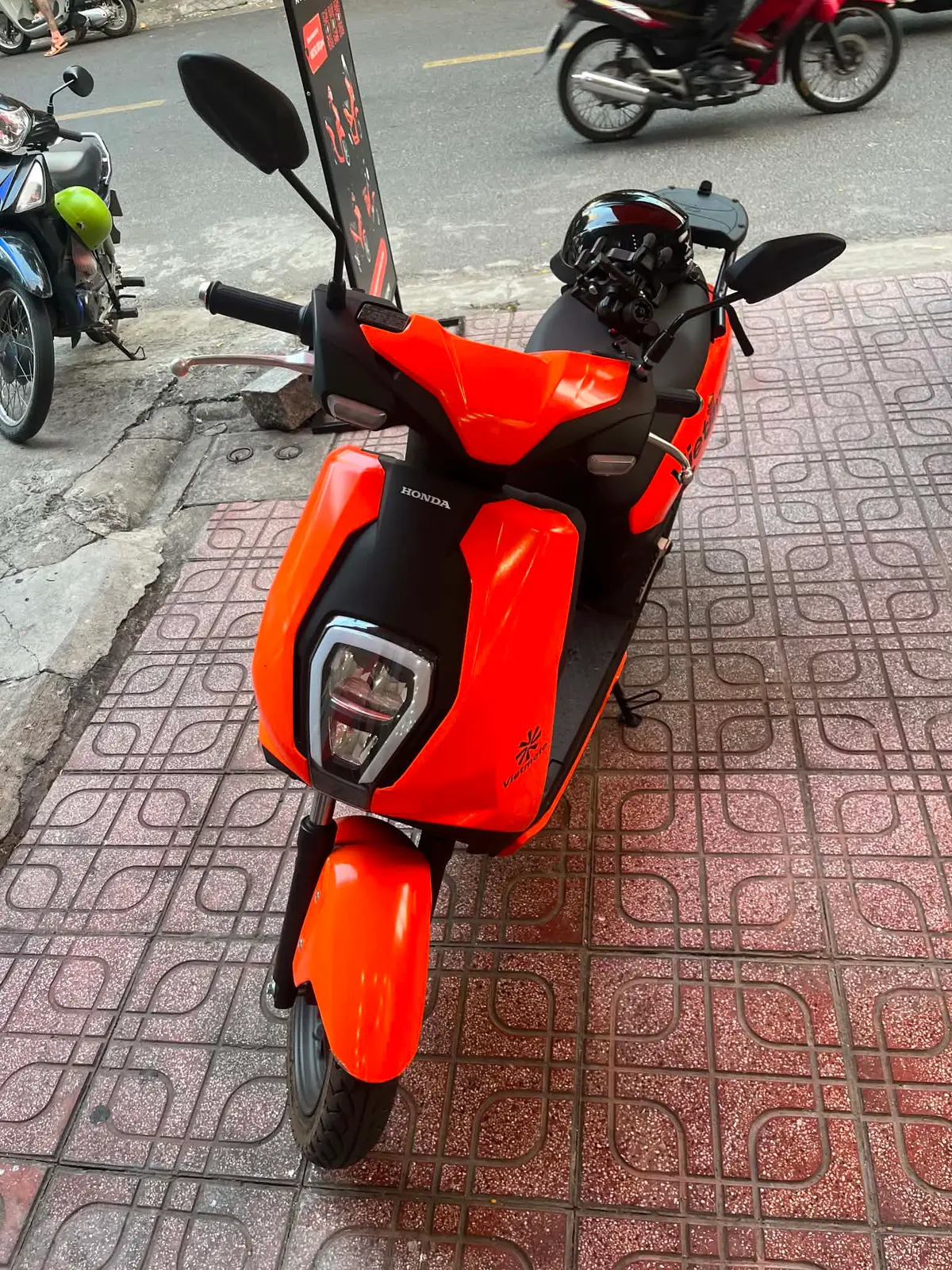 Front of an orange electric scooter with a Honda badge parked on a patterned pavement