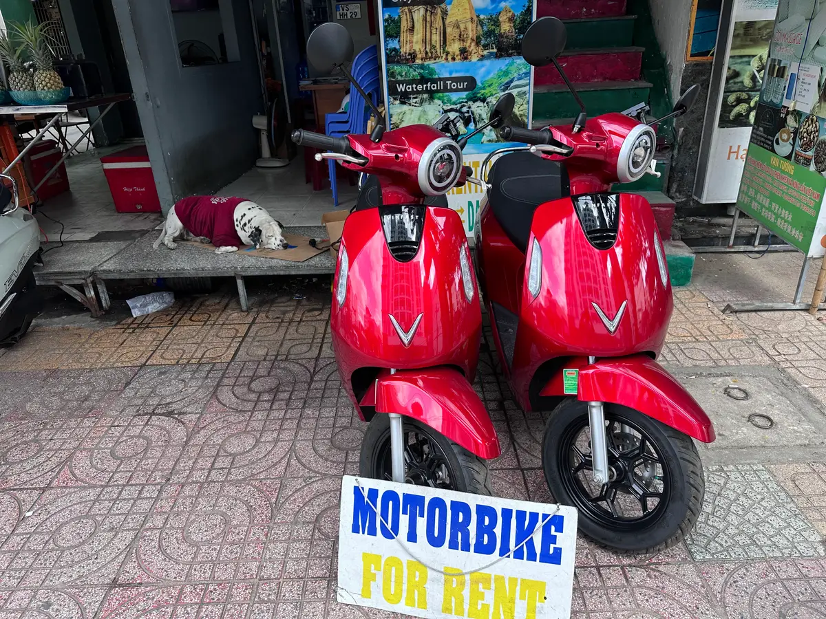 Two red electric scooters parked at a shop entrance beside a sign reading Motorbike for rent