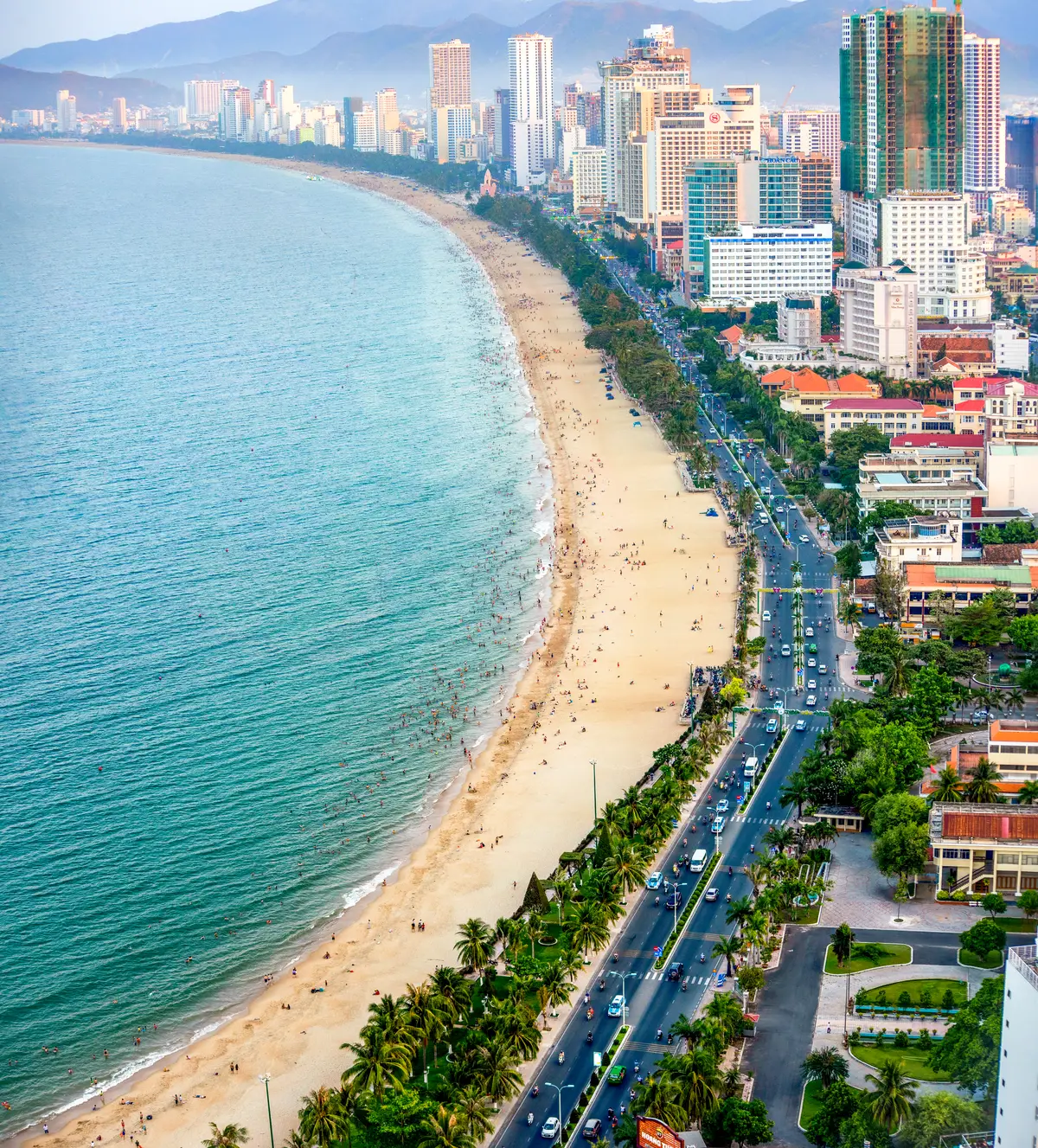Aerial view of a sandy beach, turquoise water and a wide road lined with trees and red-roofed buildings