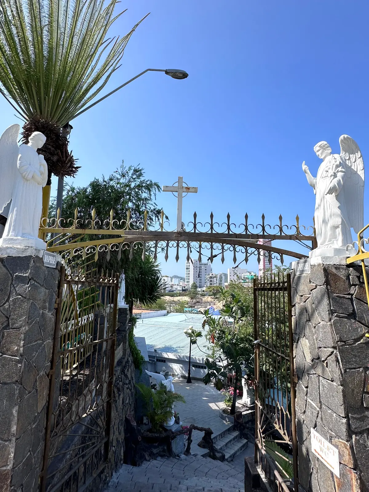 White angel statues beside a gate with a cross and city buildings behind