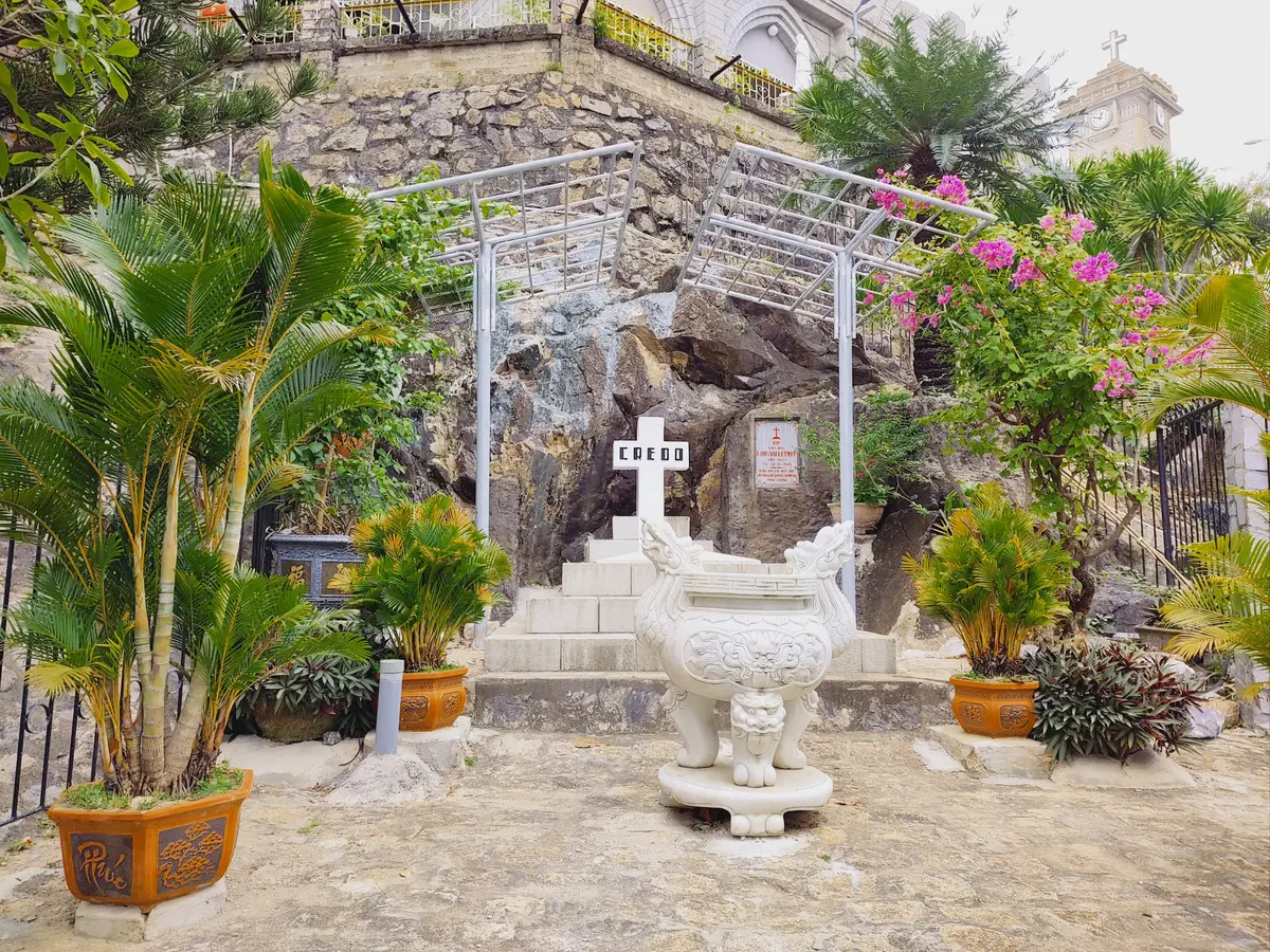 Small stone grotto shrine with a cross and potted plants beside the church