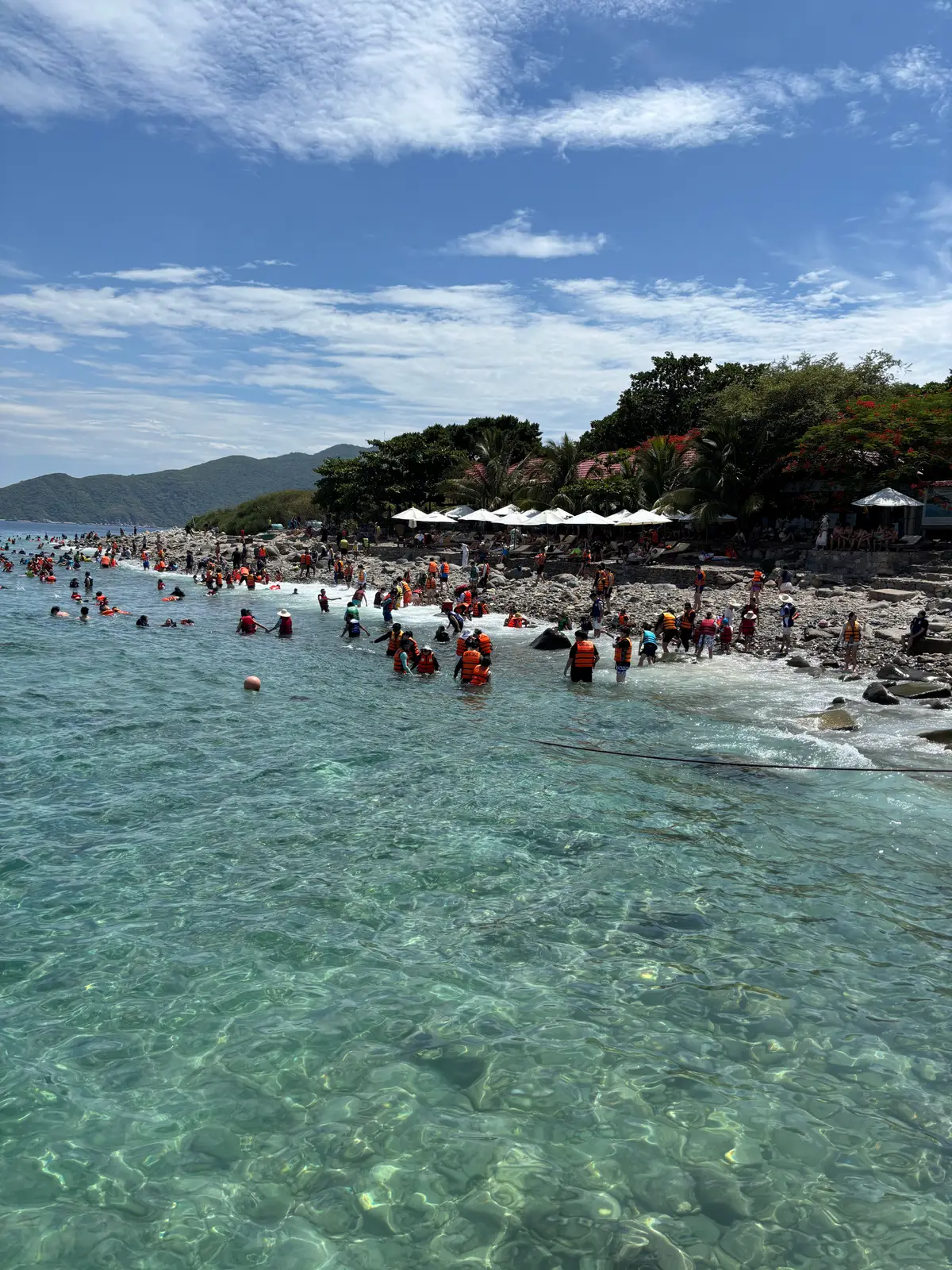 Visitors wading in shallow turquoise water along a pebble beach with white parasols