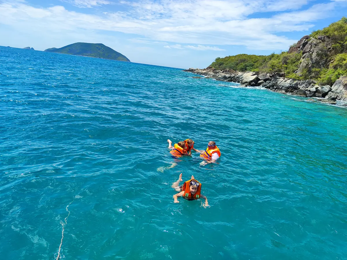 Three swimmers in orange life jackets in blue water below a green headland