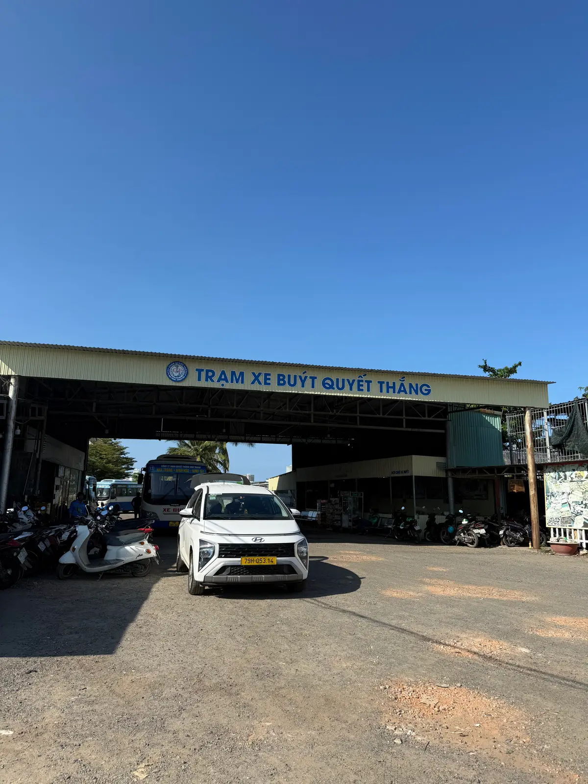 Covered entrance of the Quyet Thang bus station with a station sign, a parked bus and a white car