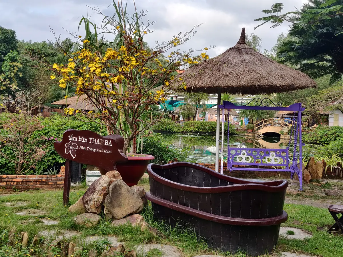 Wooden mud-bath tub in a garden beside a thatched parasol and a small signboard