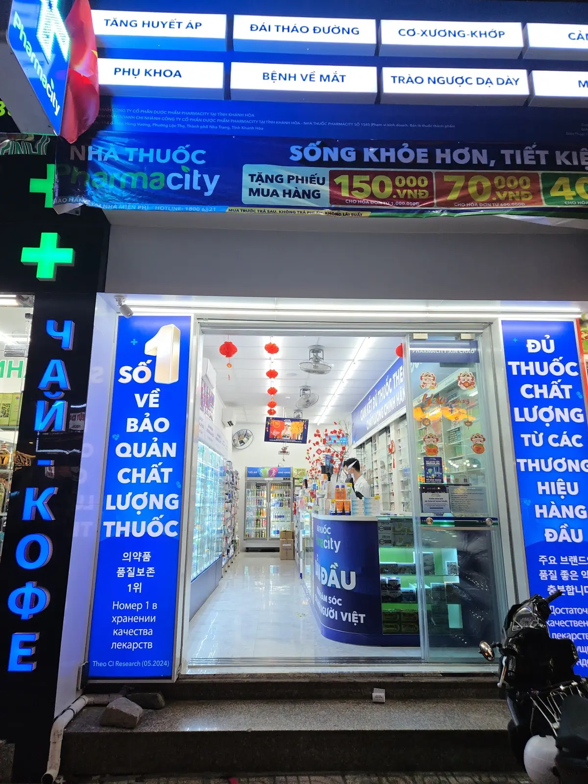 Lit pharmacy entrance with a green cross sign and multilingual shop signs
