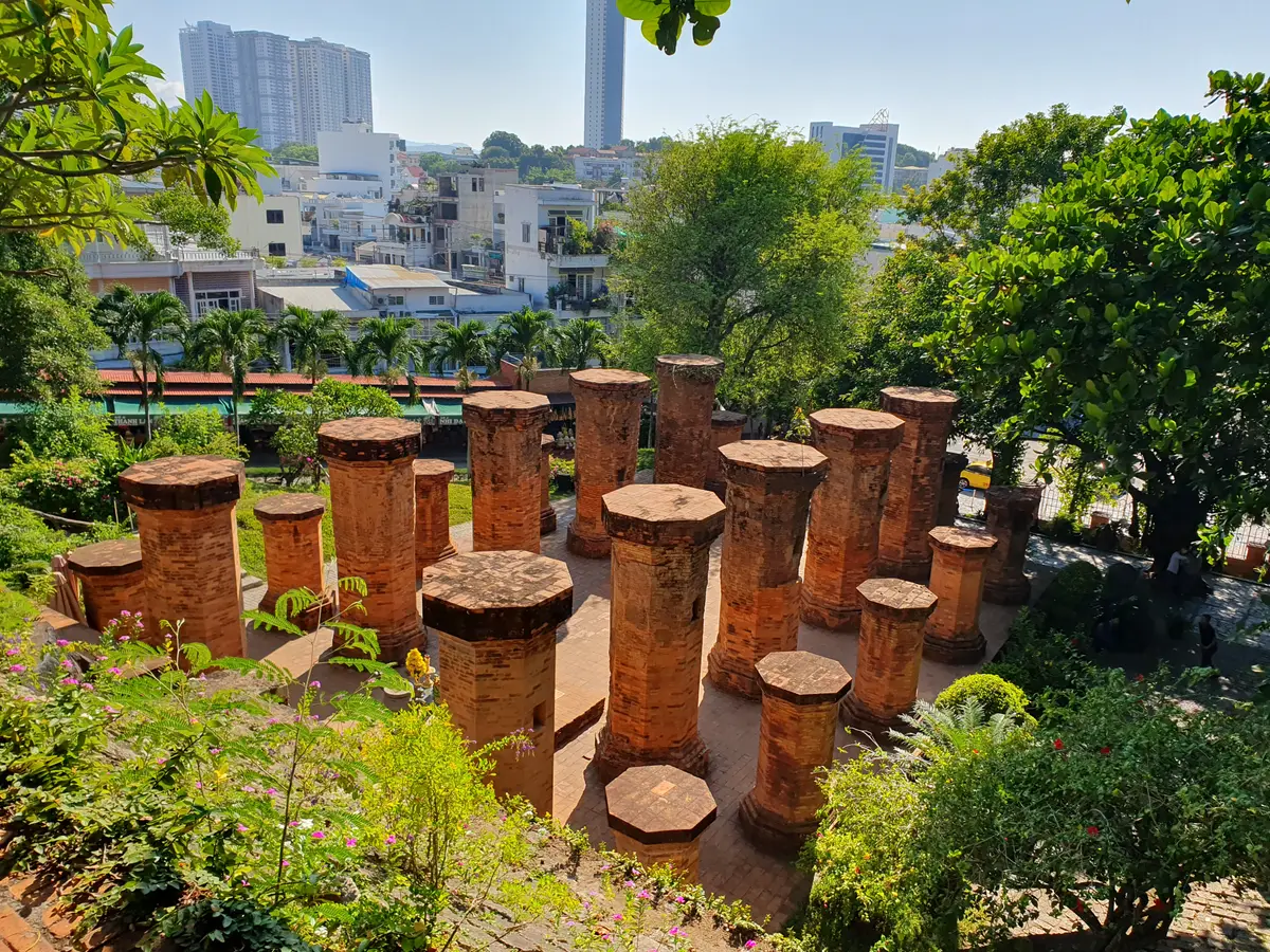 Rows of round brick pillars of the mandapa with green trees and city roofs behind