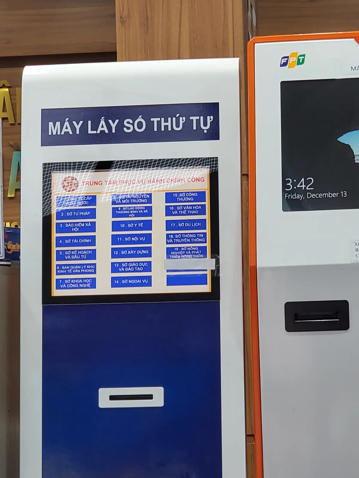 Blue queue-ticket machine with a screen listing the service windows in Vietnamese, standing against a wall beside a second kiosk