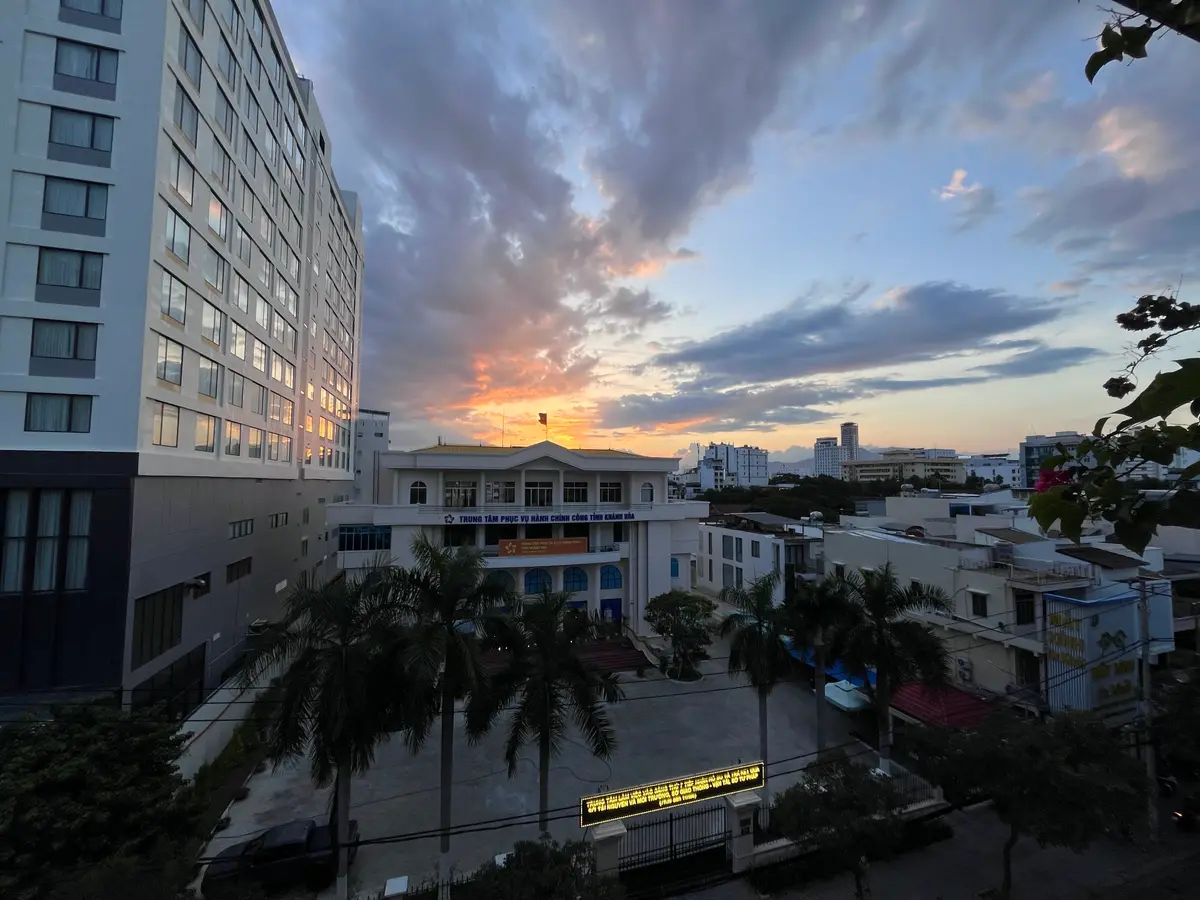 Dusk view over Nha Trang with orange clouds, a multi-storey building on the left and low government buildings and palm trees along the street below