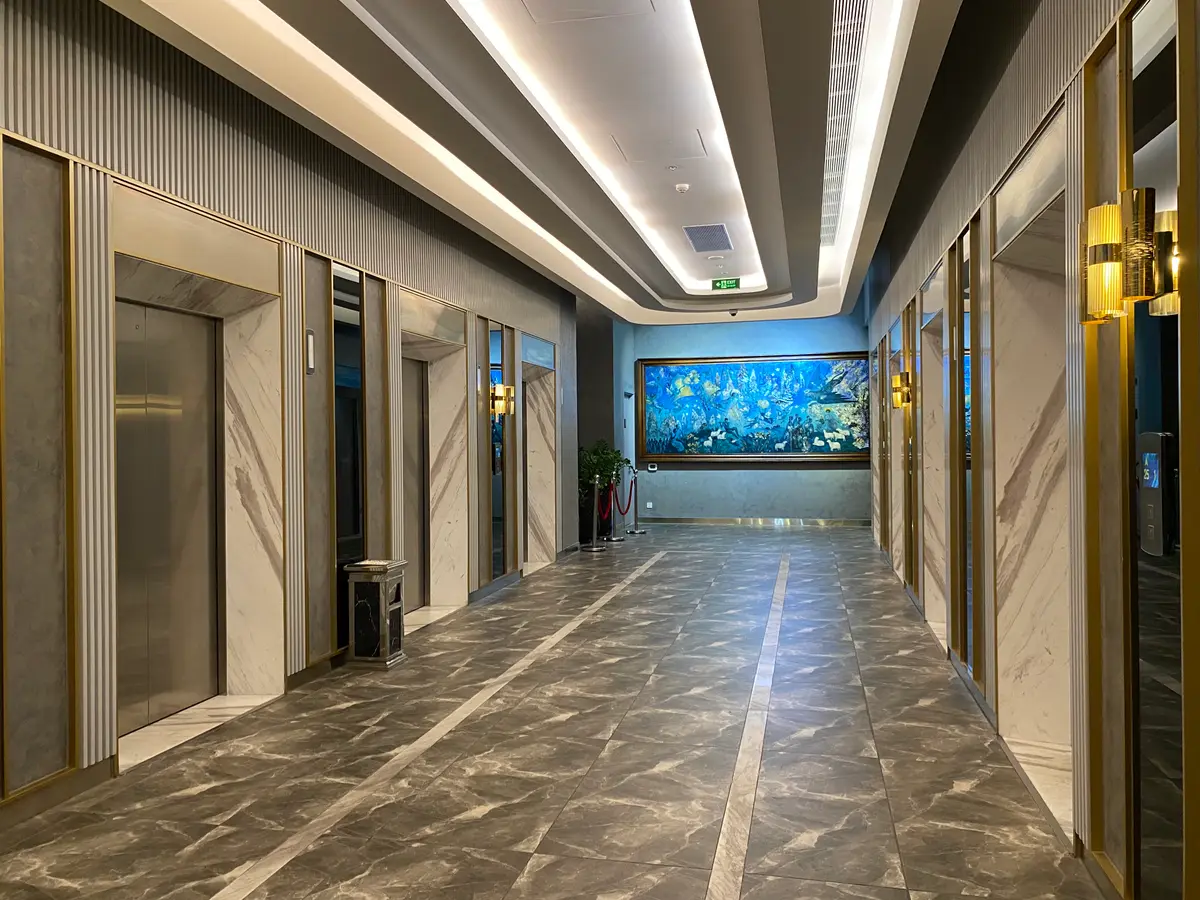 Marble-floored lift corridor in an apartment tower with closed lift doors on the left and a lit aquarium screen at the end