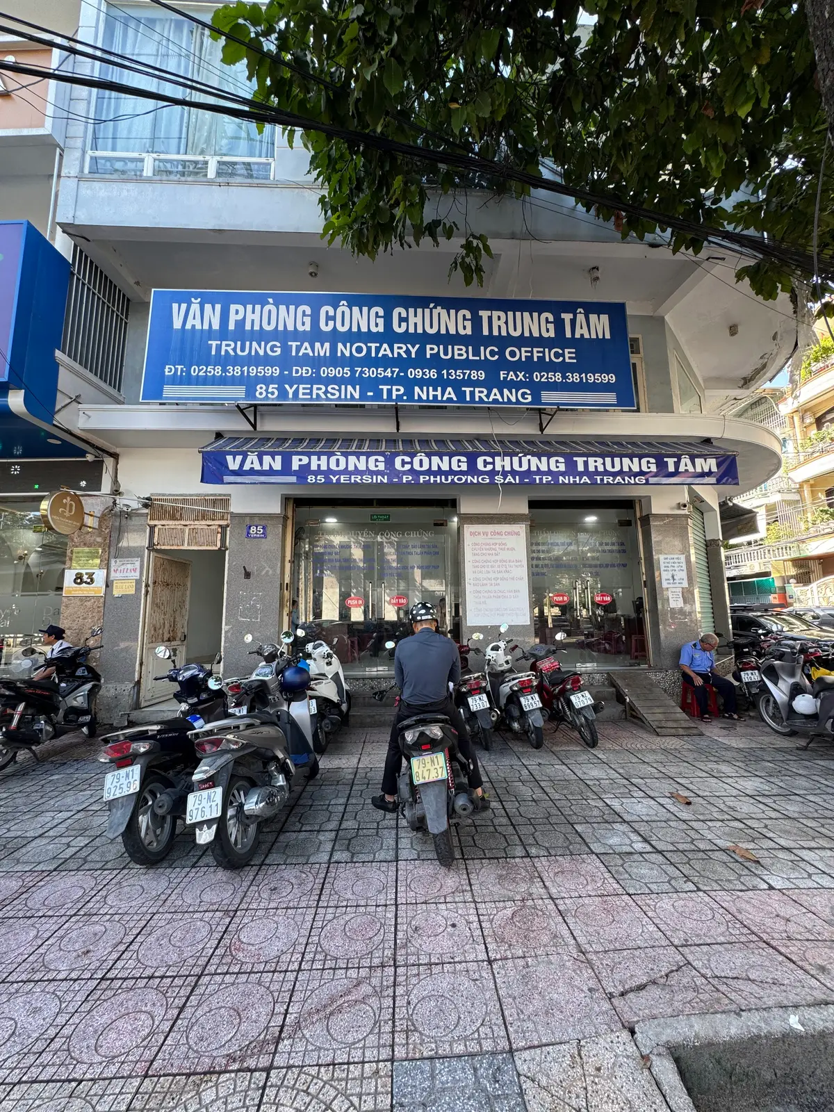 Notary office with a blue sign above the glass door and a yard full of parked motorbikes in front