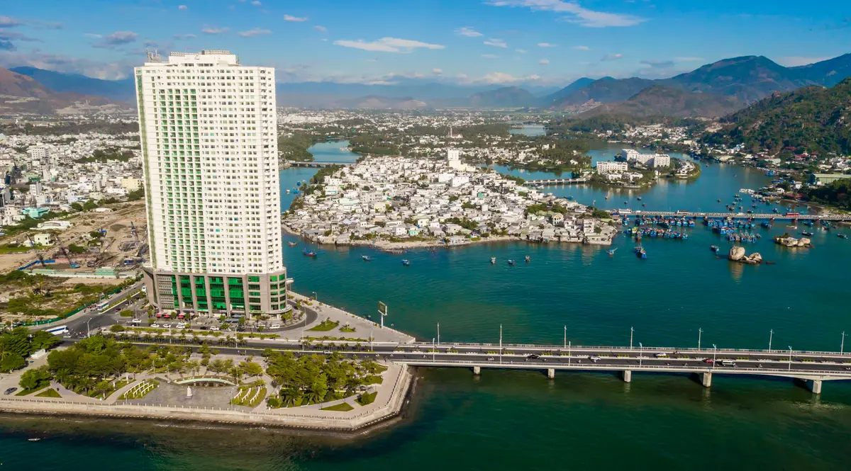 Aerial view of a white tower beside a long bridge and a bay with a town on the far shore and mountains behind