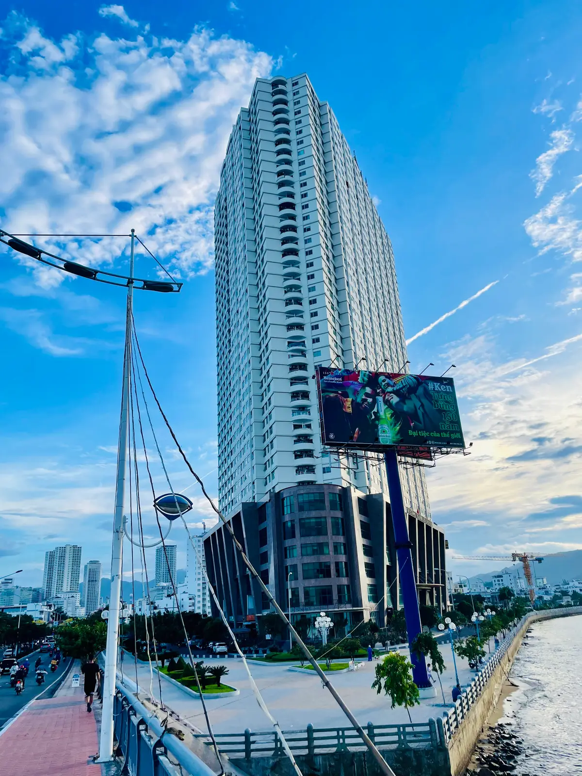 Tall apartment tower with a mast and a large screen in front of it, with city buildings below under a blue sky