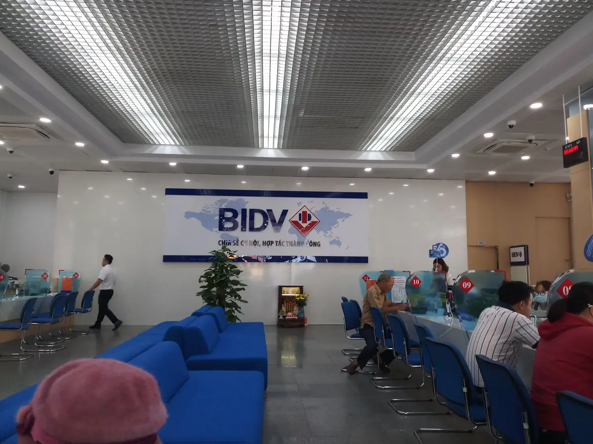 Banking hall with a blue BIDV logo on the back wall, blue seats and customers at the counters.