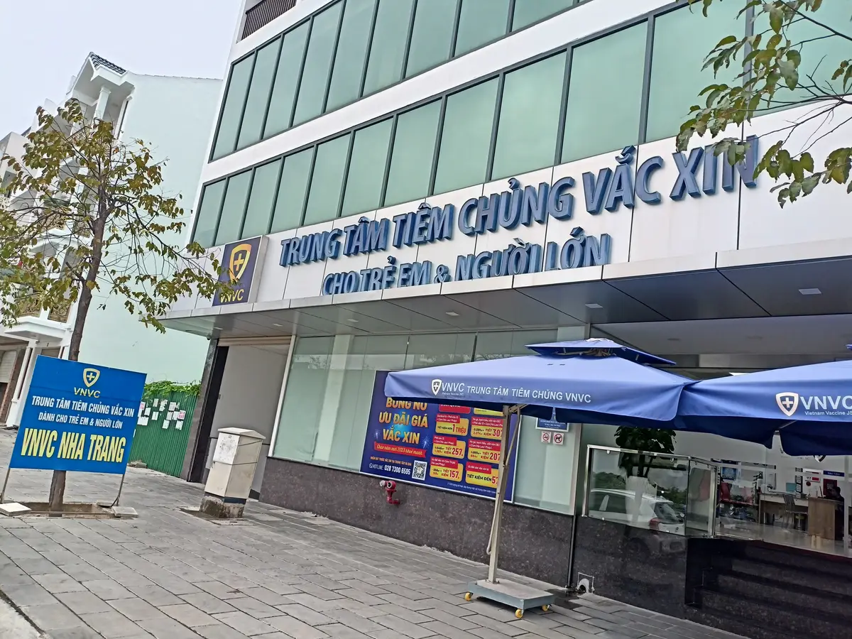 A ground-floor entrance with a blue VNVC vaccination centre sign above glass doors and a blue umbrella outside.
