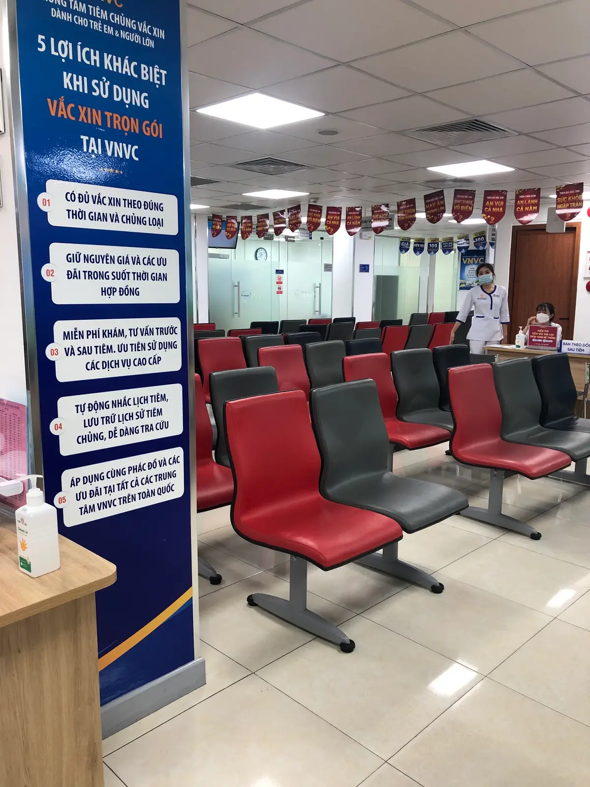 Rows of red and grey waiting chairs facing a reception counter where a nurse in a mask sits.