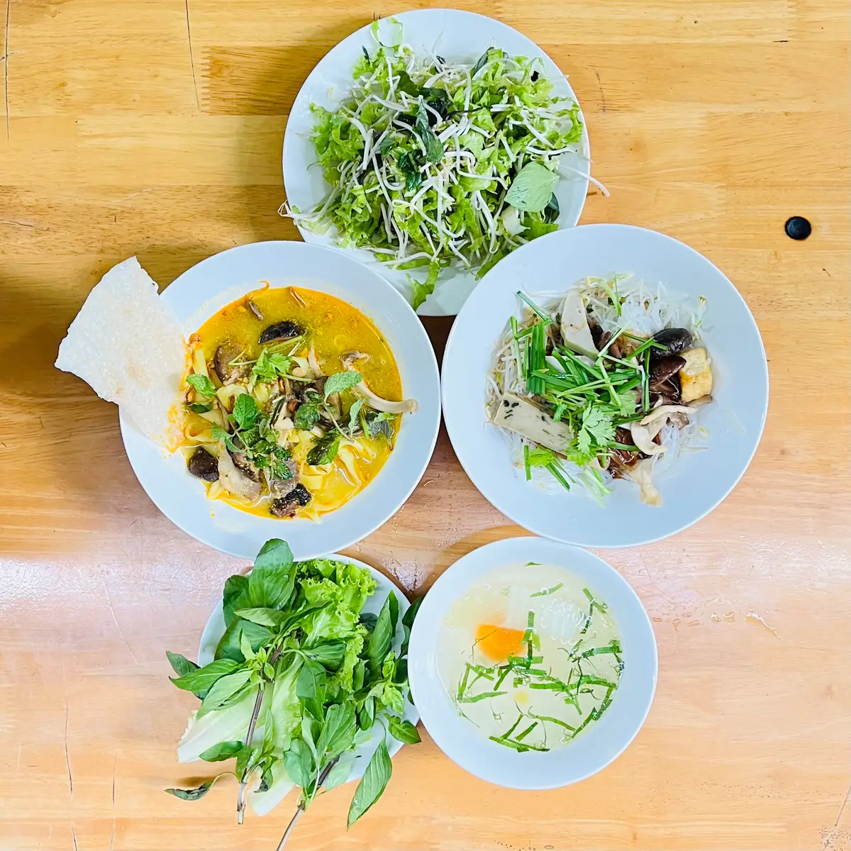 Overhead view of noodle bowls in yellow broth beside plates of fresh herbs