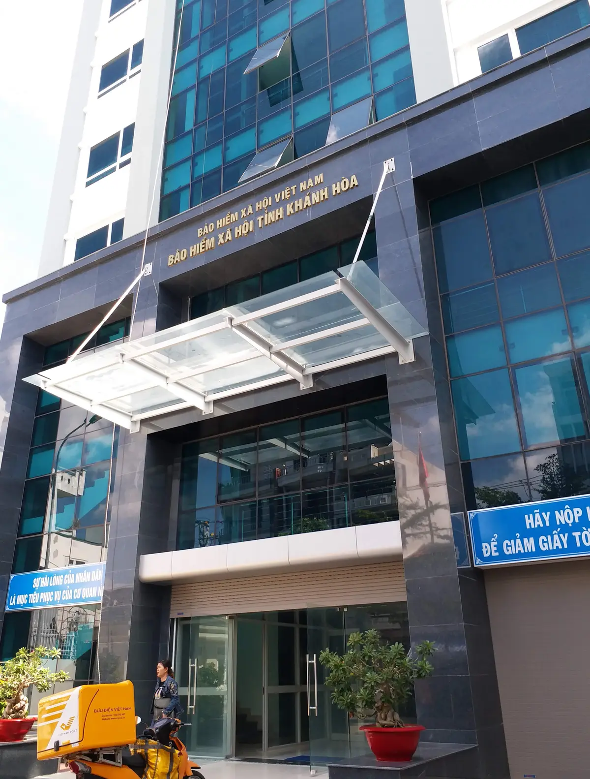 Glass facade of an office building with a canopy over the entrance and a blue sign beside it.