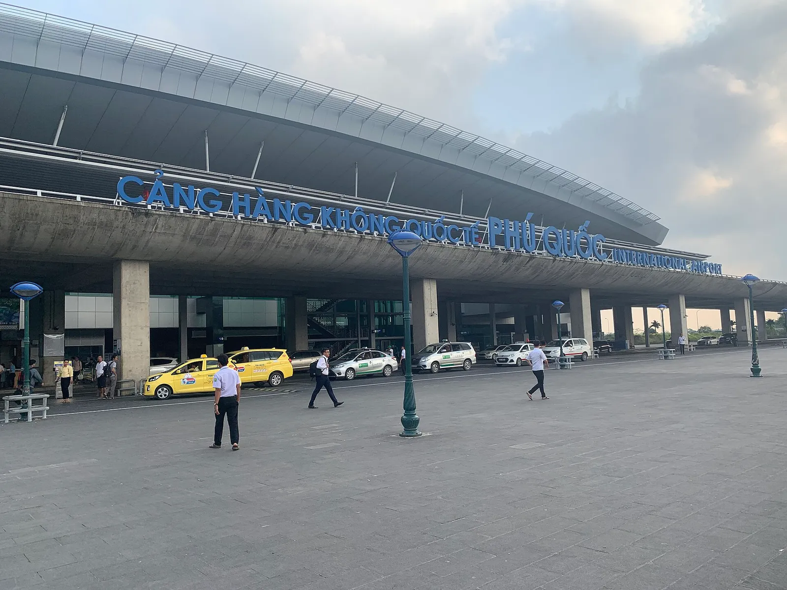 Front of an international airport terminal with a large blue sign across the roof edge, taxis parked outside and people walking in front.