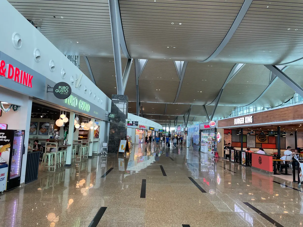 Long airport concourse with shops and a cafe on the left, wide glass roof beams overhead and travellers in the distance.