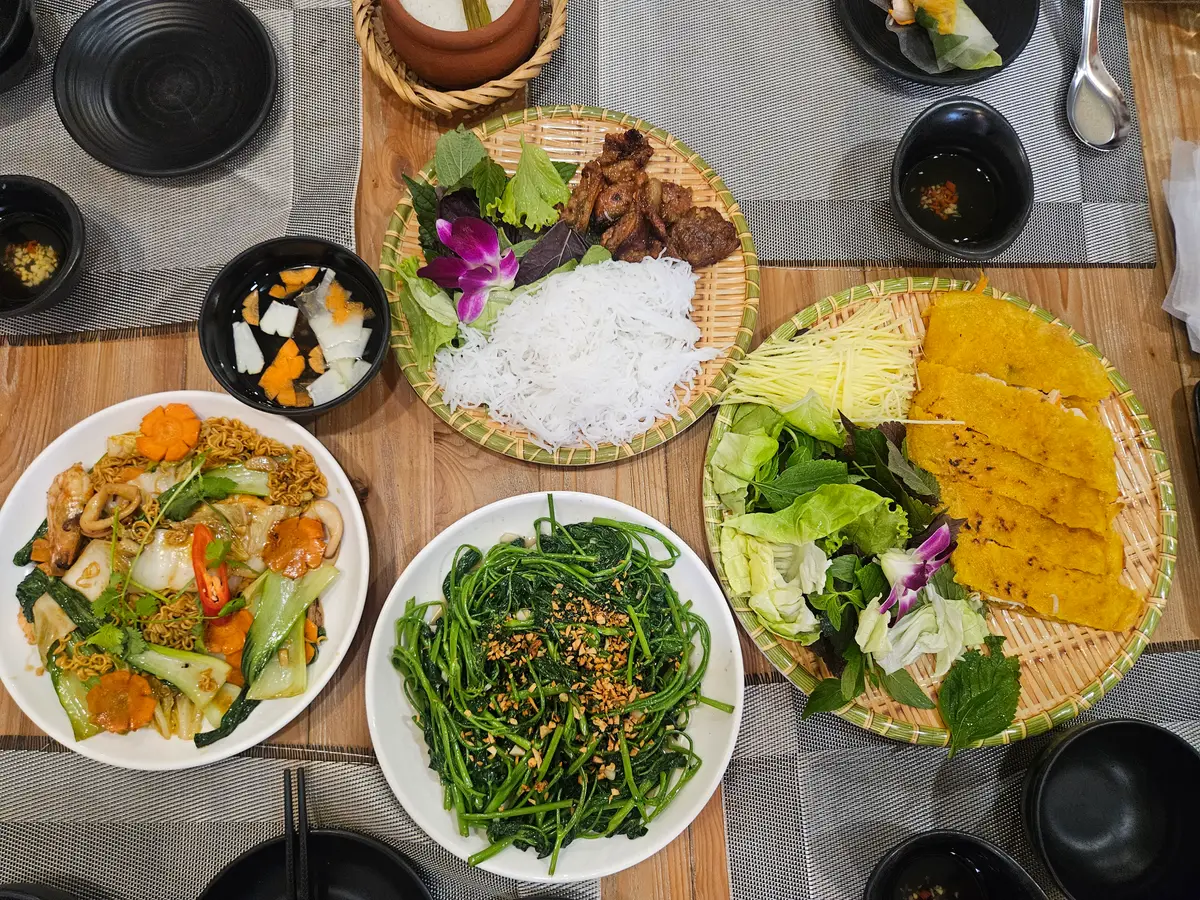 Table with a savoury pancake, rice, grilled meat, stir-fried water spinach and vegetables