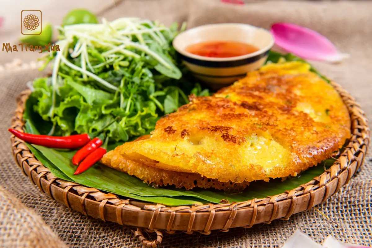 Golden crisp savoury pancake on a woven tray with herbs, chilli and dipping sauce